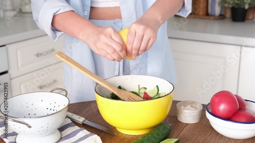 Person squeezing lemon over fresh salad in bright home kitchen with yellow mixing bowl, wooden spoon, tomatoes, cucumber, colander, and soft daylight on wooden table