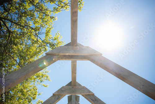 Photos Low angle view of a timber roof truss and ridge beam in a garden, spring leaves nearby, strong sun flare, wide angle composition, warm wood tones, clean lines
