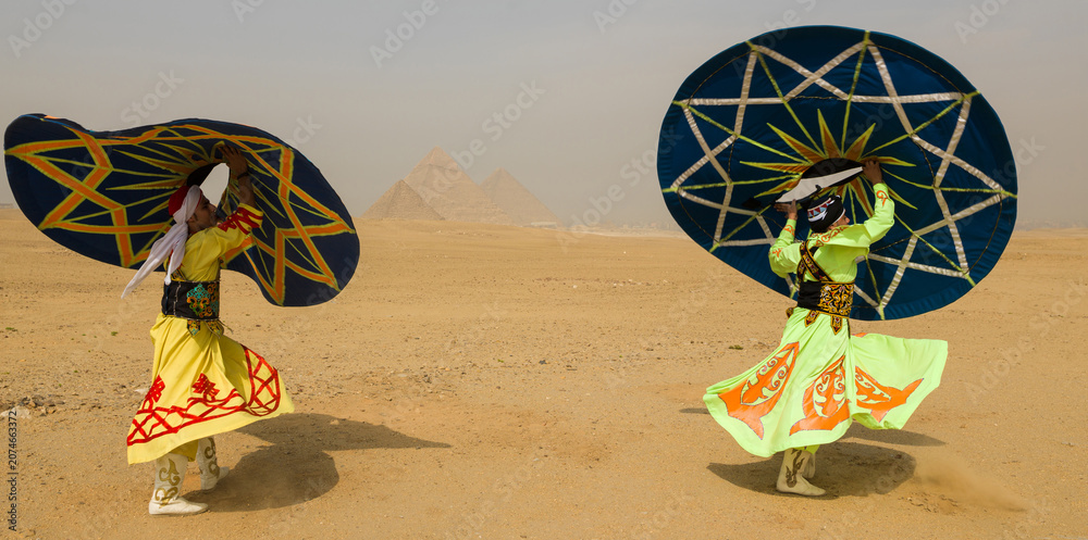 Fototapeta premium Two vibrant Tanoura folk dancers spinning colorful star patterned skirts overhead in the desert before the Giza pyramids in Egypt ideal for tourism