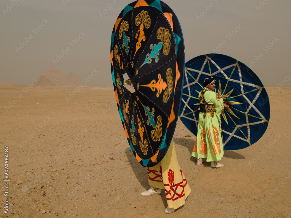 Fototapeta premium Vibrant Egyptian tanoura folk dancers carrying their colorful spinning Sufi skirts across the desert sand with the Giza pyramids behind ideal for