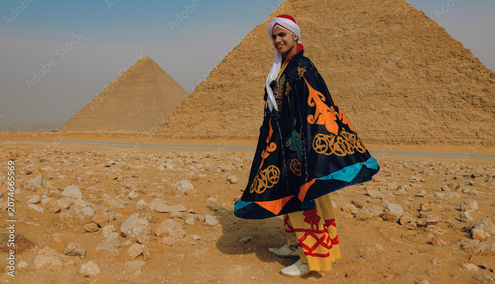 Fototapeta premium Joyful young Egyptian tanoura folk dancer twirling a colorful ornate cape in front of the Giza pyramids ideal for Egypt tourism cultural festival and
