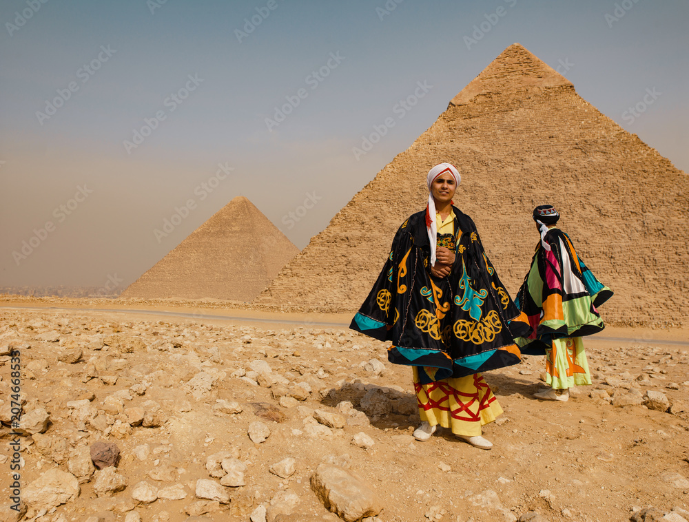 Fototapeta premium Festive Egyptian folk dancer in a colorful embroidered tanoura costume and turban standing proudly before the Giza pyramids in the sunny desert ideal