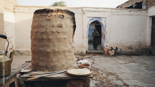 Mud oven stands in a stone courtyard as a woman enters a traditional home through a small arched doorway nearby.