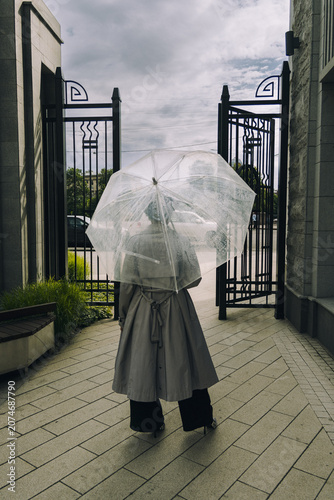 Woman walking toward open black metal gates. She holds transparent umbrella viewed from behind. Overcast sky above paved pathway flanked by stone walls. Atmospheric, solitary, mysterious urban scene.