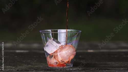 Refreshing red beverage pouring into a clear plastic cup filled with ice cubes. The cup sits on a dark wooden picnic table outdoors in slow motion