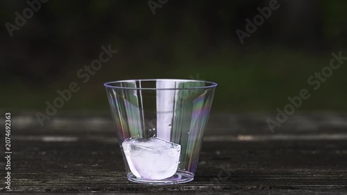 Slow motion close-up view of a person's hand placing ice cubes into a clear plastic cup resting on an outdoor wooden table for a refreshing drink
