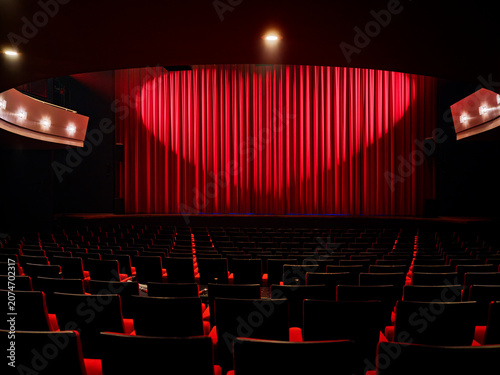Red stage curtain and empty seating rows in a theater auditorium with warm stage lighting, a performing arts hall interior