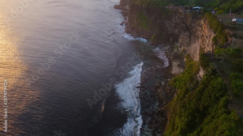 Aerial perspective of powerful white sea foam waves washing over coastal rocks beneath dramatic sunlit cliffs. Stunning natural contrast.