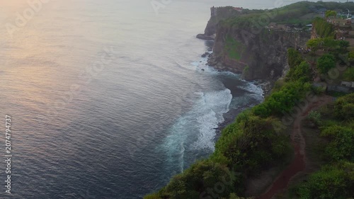 Aerial perspective of powerful white sea foam waves washing over coastal rocks beneath dramatic sunlit cliffs. Stunning natural contrast