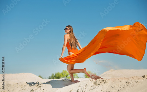 A beautiful young woman with a beautiful body and skin in a swimsuit and glasses in motion on the sand against the sky