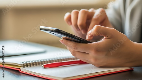 A person uses a smartphone on a table with a notebook and pen.