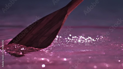 Close-up of a wooden paddle cutting through shimmering violet water creating ripples in a cinematic atmosphere
