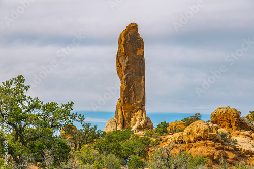 Photos The Tower of Babel, a tall freestanding sandstone pillar, rises above green junipers against a soft cloudy sky, Arches National Park, Utah