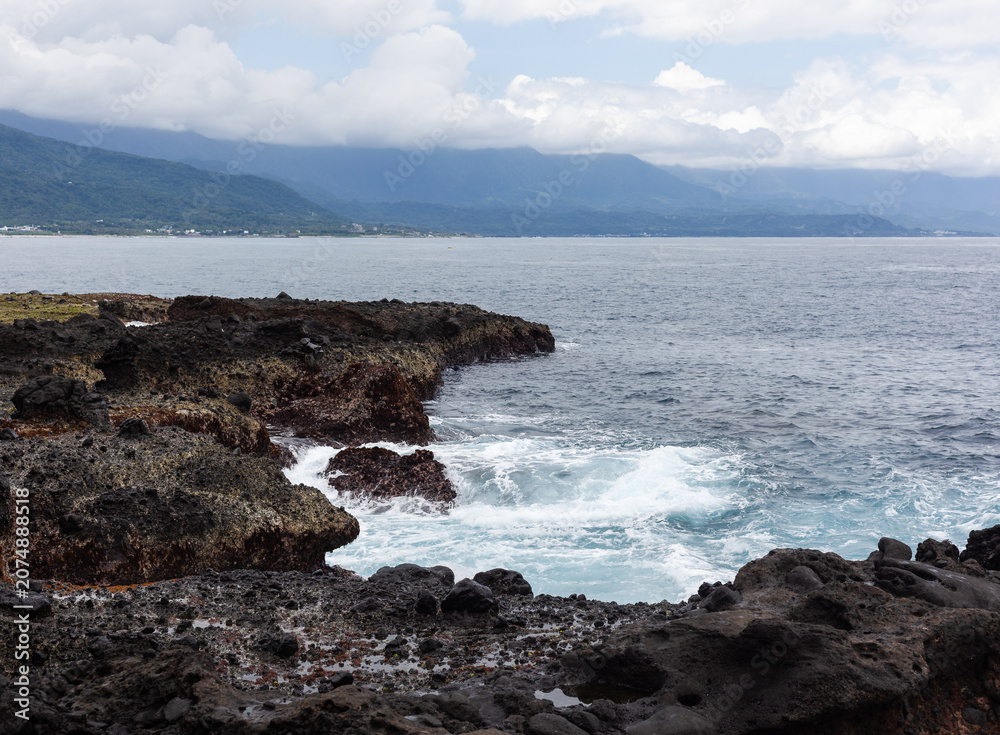 Fototapeta premium Sanxiantai rocky coastline with waves crashing. Distant mountains under cloudy sky. Taiwan's scenic eastern shore.