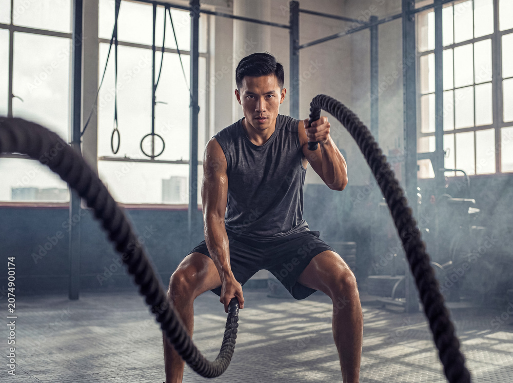 Obraz premium Muscular Asian man vigorously training with heavy battle ropes in a well-equipped, industrial-style fitness gym.
