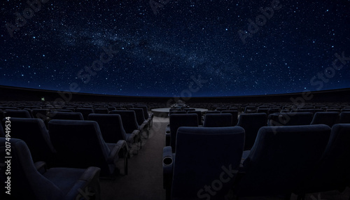Empty planetarium theater with rows of dark seats facing a large domed ceiling displaying a realistic night sky with the bright milky way galaxy