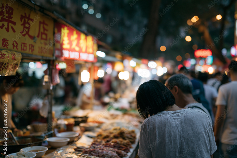 Fototapeta premium Night market glows with warm, inviting lights. Shoppers browse stalls filled with fresh foods. Woman leans in, curious about the offerings. Vibrant signs glow softly against the night