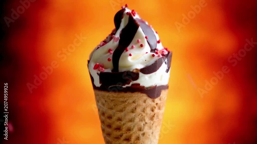 Slow motion shot of red sprinkles falling on a delicious vanilla soft serve ice cream cone with chocolate syrup. Sweet summer dessert concept against a vibrant orange background