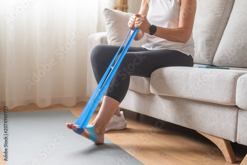 Bild auf Leinwand Woman sitting on a sofa performing foot rehabilitation exercises with a blue res