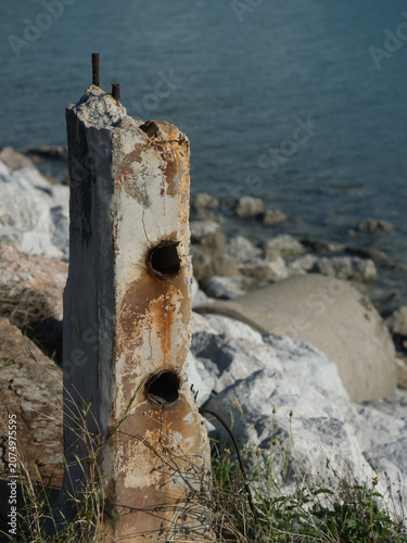 Rusted iron bars are attached to weathered concrete pillars along the seashore the exposed metal rebar is corroded by the salty air set against a backdrop of a green sea.