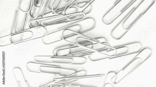 Top-down view of silver metal paper clips scattered on a white background. Slow rotating camera movement highlighting office supplies and stationery items