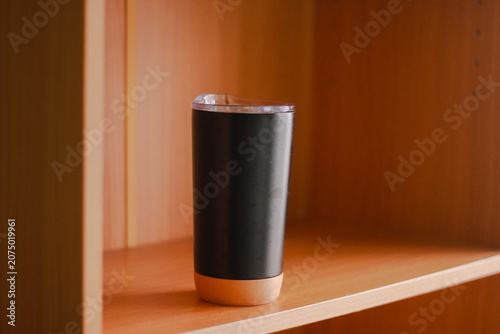 A sleek black insulated travel tumbler with a transparent lid stands on a wooden shelf in warm natural light. Its minimalist design and modern aesthetic make it ideal for coffee, tea, and everyday hyd
