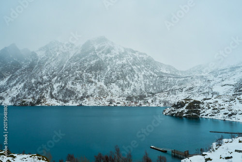 Winter Road Trip in Lofoten: Small Village Nestled Under Majestic Snow-Covered Mountains by a Dark Fjord, Moody Arctic Landscape and Scenic Wilderness in Norway.