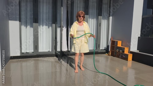 Barefoot woman is washing tiles with green garden hose on open patio. Water stream cleans floor under bright sunshine, showing cozy home routine, high pressure and relaxed summer lifestyle.