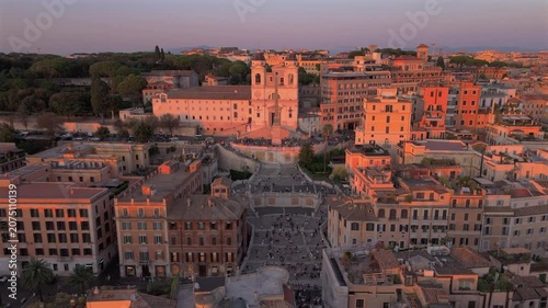 Aerial drone shot in Rome Italy captures the iconic Spanish Steps and Trinita dei Monti Church bathed in warm golden hour light with historic terracotta rooftops and tourists gathering below.