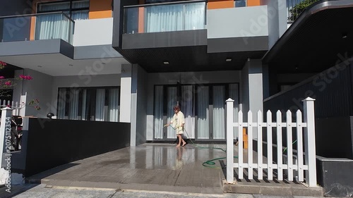 Woman does courtyard maintenance by washing driveway with hose near modern townhouse. Clean outdoor area, white fence and bright sunlight reflect high living standards in suburban neighborhood.