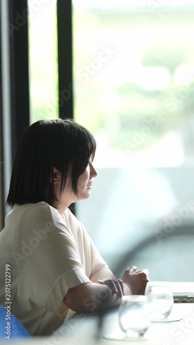 Soft light fills a quiet cafe corner. Woman sits, gazing out the window. Her hands move as she listens attentively. Cups rest on the table beside her. Peaceful, thoughtful, and calm