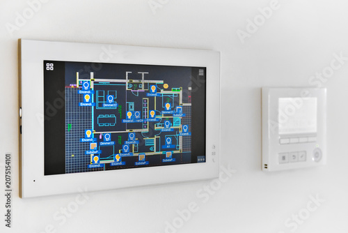 A smart home automation system is mounted on white wall, displaying digital floor plan with various controls, closeup