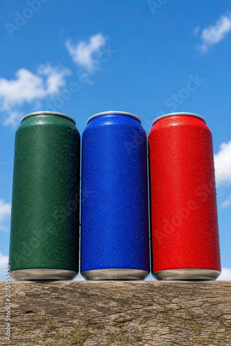 Three Blank Colored Aluminum Beverage Cans with Water Drops Standing in a Row on Wooden Surface Against Blue Sky and Clouds, Cold Soda and Beer Concept