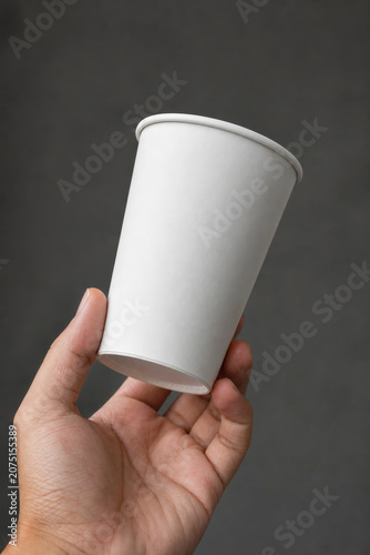 Close-up of a person holding a blank white paper coffee cup against a dark, textured grey background, takeaway beverage mock-up concept