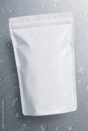 Top view of a blank white zipper pouch bag covered in fresh water droplets, lying on a metallic gray background, wet packaging mockup