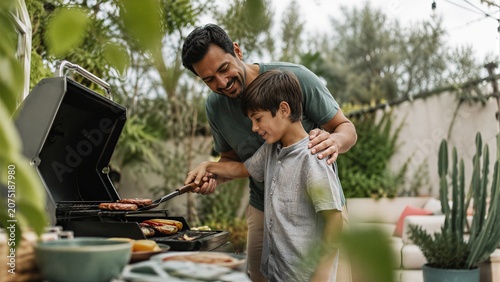 Happy father grilling burgers with his son in the garden. Man helping boy cook meat on barbecue. Family bonding and summer BBQ concept for Fathers Day celebration
