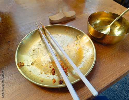 empty golden plate with chopsticks and a small bowl on a wooden table after a meal. The leftover food pieces and dining utensils create a casual restaurant atmosphere with warm lighting and a cozy