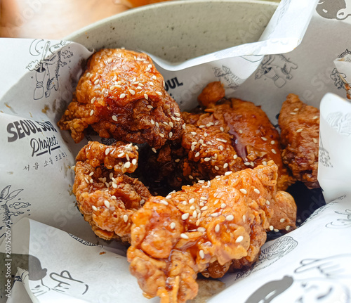 Crispy Korean fried chicken pieces coated with a sweet and spicy sauce and topped with sesame seeds. The chicken is served in a paper box, showing a delicious and appetizing street food style.
