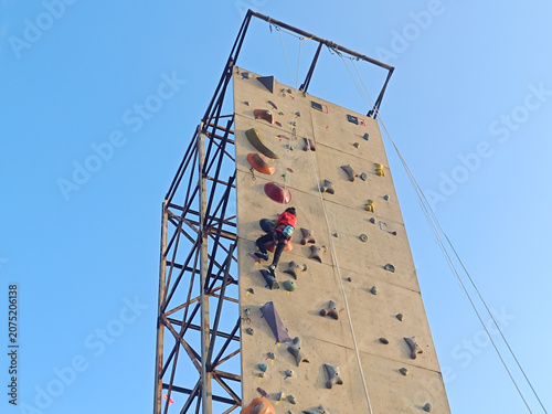 young person is climbing an outdoor rock climbing wall while secured with a safety rope. The tall climbing wall has colorful climbing holds and stands against a clear blue sky