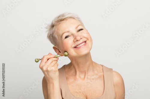 Gorgeous woman of 60s massaging face skin using natural facial stone trendy jade green quartz roller