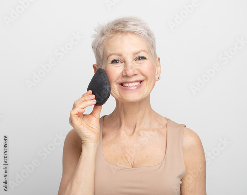 Charming elderly woman with short hair use sponge remove make up on light grey background.