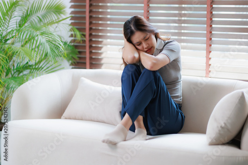 Vászonkép Sad asian woman feeling down and distressed, hugging knees on a sofa at home, ba