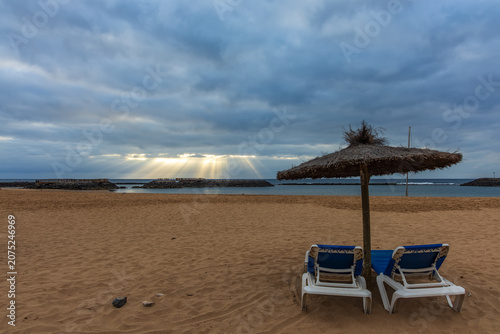Schilderij op canvas Stunning crepuscular sunbeams over Playa de la Guirra beach with empty loungers,