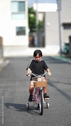 Child rides bicycle down quiet street. Child smiles, enjoying simple joy of motion. Basket holds small items, adding charm to ride. Sunlight casts soft shadows on asphalt