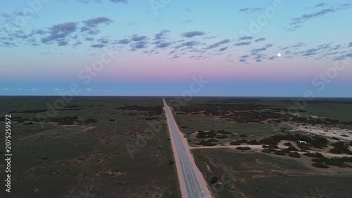 Aerial drone shot flying forward above the endless straight road on the Nullarbor Plain with a white moon over a blue‑pink‑purple dawn sky