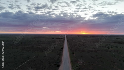 Aerial drone shot flying forward above the endless straight road on the Nullarbor Plain with a red sunrise and clouds glowing in the morning light