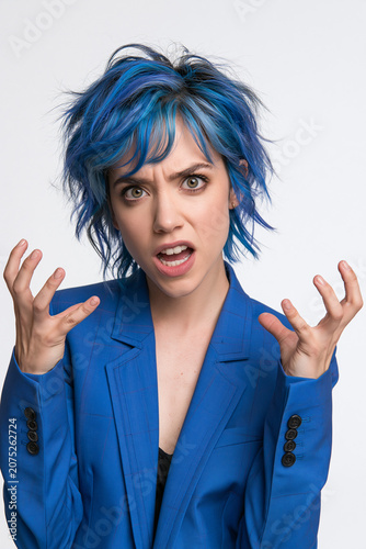 Aggressive and Frustrated Young Woman with Bright Blue Short Hair Wearing a Matching Blue Blazer Shouting and Gesturing with Her Hands Against a Clean White Background Studio Portrait.