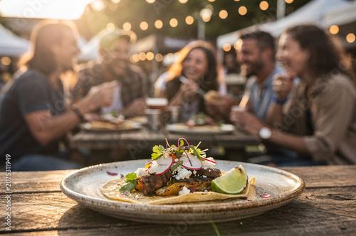Delicious gourmet street food taco served on a plate at a lively outdoor festival with blurred friends dining in the background