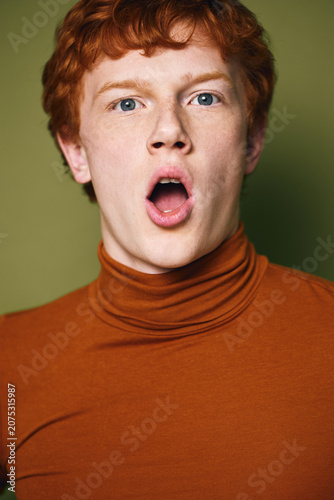 Canvastavla Young man with red hair and blue eyes wearing orange turtleneck looking surprised or shocked with mouth open on green background for emotion and expression concept