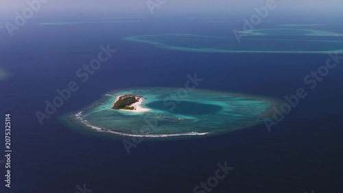 Aerial pan shows a ring shaped atoll in Maldives, a sandy palm islet in a turquoise lagoon, white surf on reef, dark ocean beyond, faint atolls on horizon in hazy daylight.